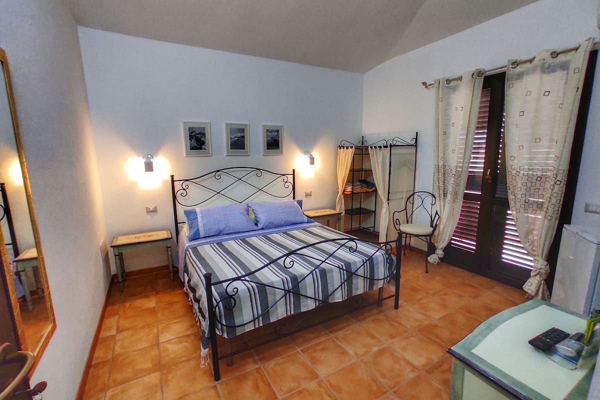 A bedroom with a bed, a chair, a small table, a nightstand, a window with curtains, and a wall-mounted shelf.