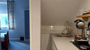 Uma cozinha com forno embutido, pia e bancada. Há uma janela com cortinas e uma porta que dá para outro cômodo.