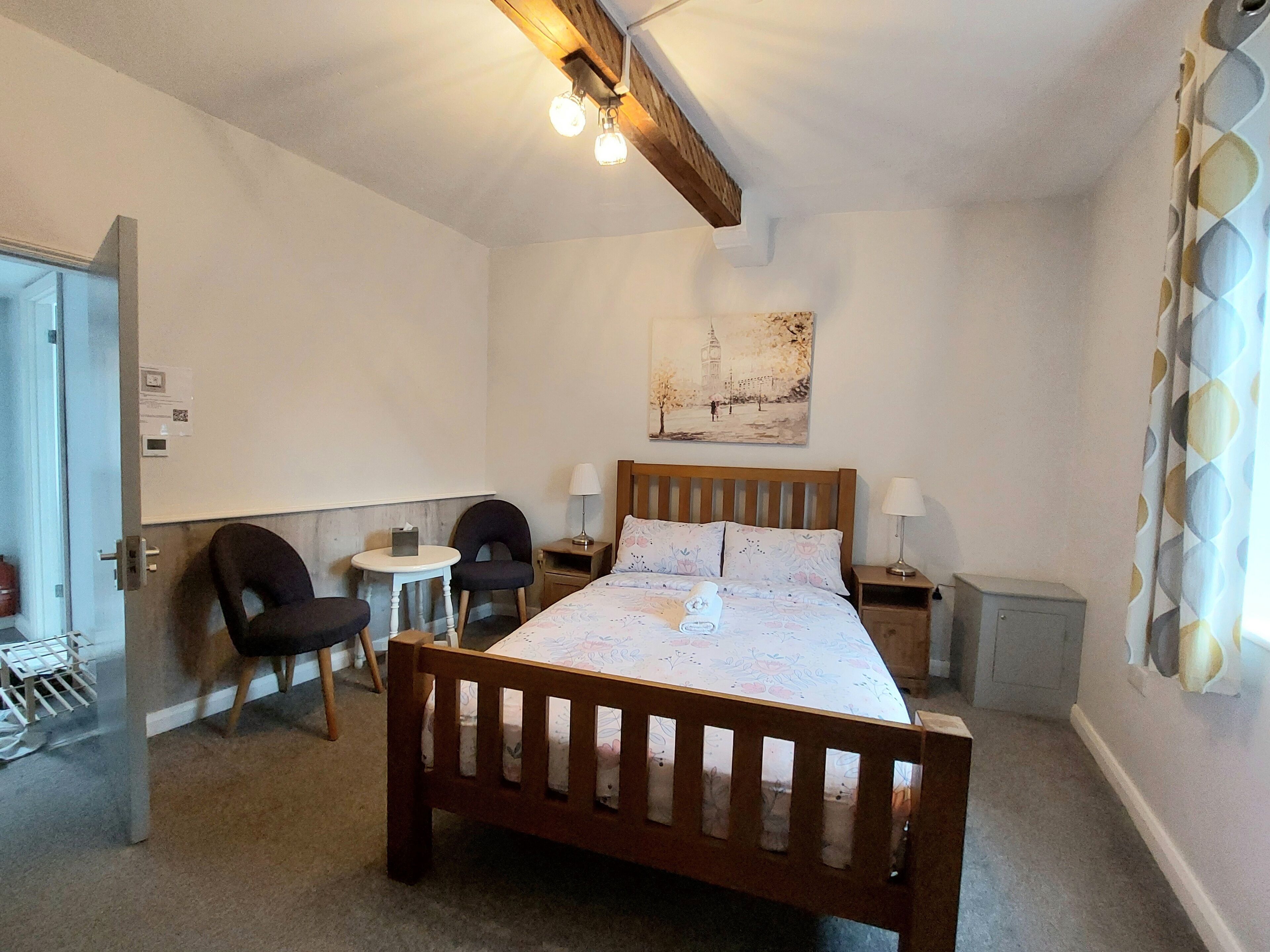A bedroom with a wooden bed, two bedside tables with lamps, a chair, a small round table, and a wall-mounted artwork.