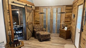 A cozy room with wooden walls, a dining table with chairs, a patterned armchair, a small wooden dresser, and a lamp. - Mine Lick Creek Resort 128 (Baxter)