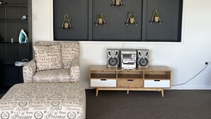 A living room with a dark wall, a large framed picture of plants, a patterned armchair, a wooden side table with speakers, and a small cabinet. - Cosy comfortable unit with city views (Napier)