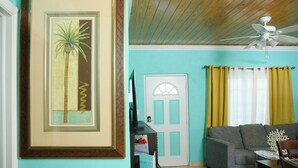 A living room with a teal wall, a framed picture of a palm tree, a grey sofa, a ceiling fan, and a door with yellow curtains. - Complete Villa Down stairs, 2 Bedroom (Steventon)