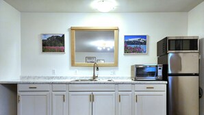 A kitchen with white cabinets, a stainless steel refrigerator, a microwave, and a sink. There are framed pictures on the wall above the sink. - Fireweed 2 Bedroom by airport; lakes, trails, fishing, skiing, hiking! (Anchorage)