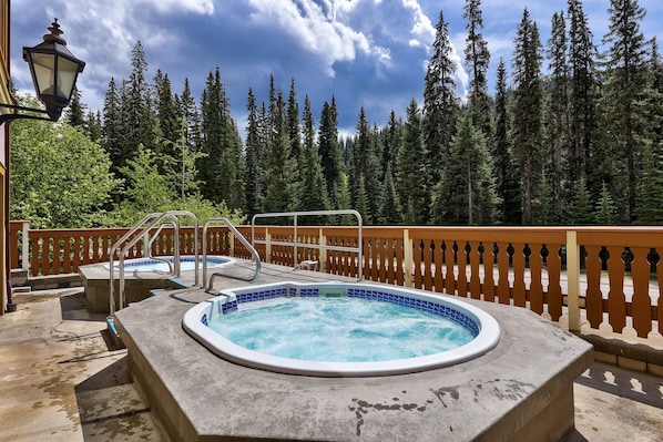 A hot tub surrounded by a wooden deck with a railing, set against a backdrop of tall evergreen trees. - Hearthstone Lodge #407 (Sun Peaks)