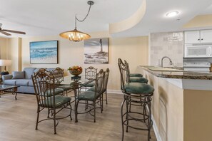 A modern kitchen with a central island, a dining area with a glass table and metal chairs, and a living room with a sofa and a painting. - Close to beach, beautiful marsh views, fitness center, indoor/outdoor pools, hot tub (North Myrtle Beach)