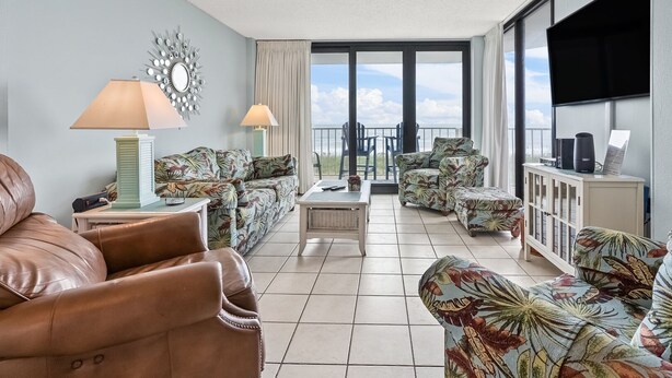 Een ruime woonkamer met een leren bank, fauteuils met bloemenprint, een salontafel en een tv op een kast.