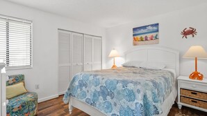 Een slaapkamer met een bed, nachtkastjes, een stoel, een kledingkast en een schilderij met een strandthema aan de muur.