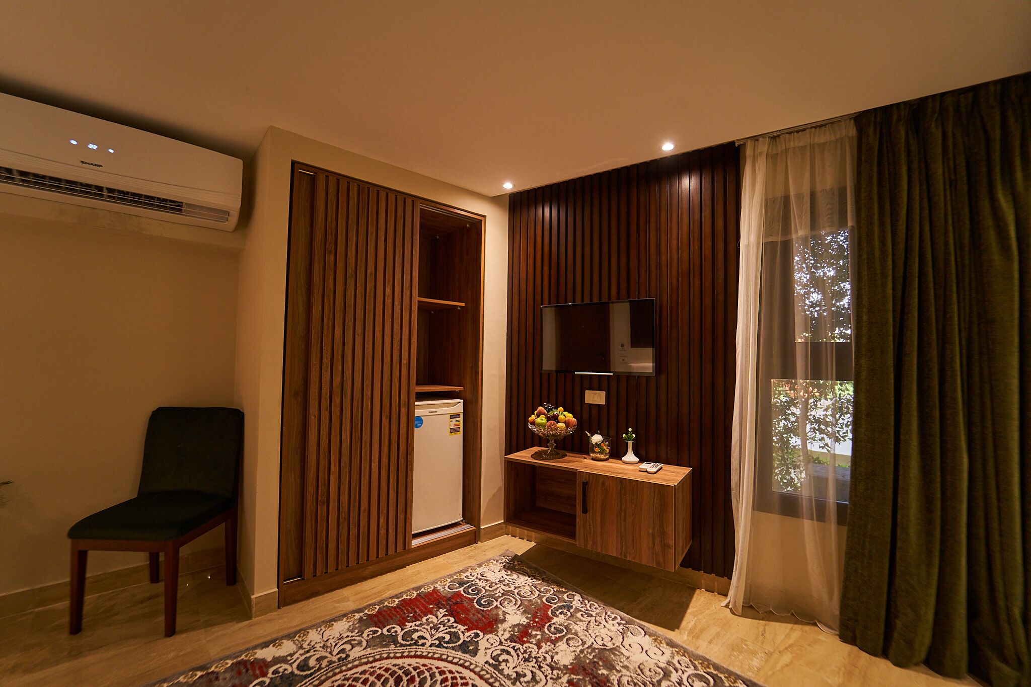 A hotel room with a wooden wardrobe, a small desk with a vase and fruit bowl, a chair, and a large window with curtains.