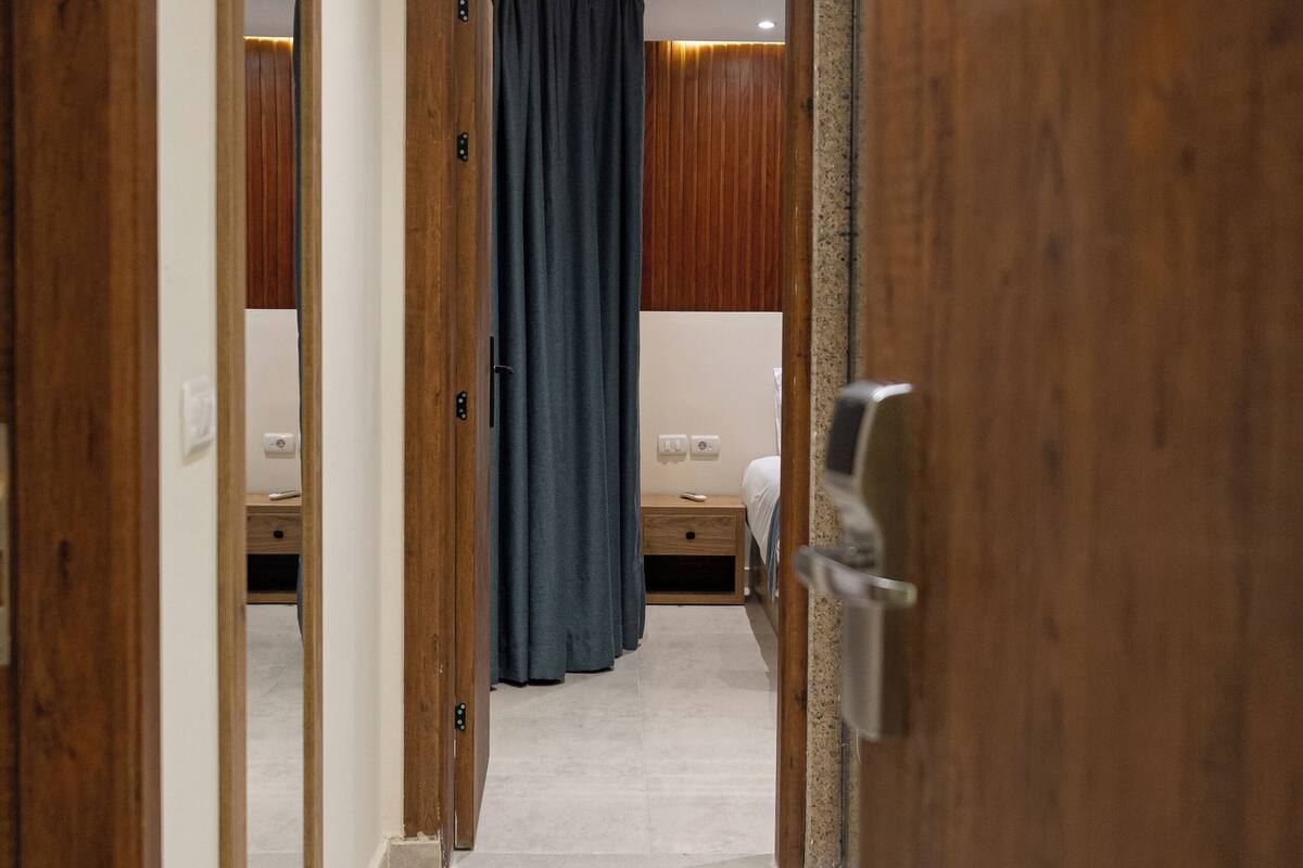 Un pasillo con puertas de madera, una cortina oscura y una cama con ropa de cama blanca al fondo.