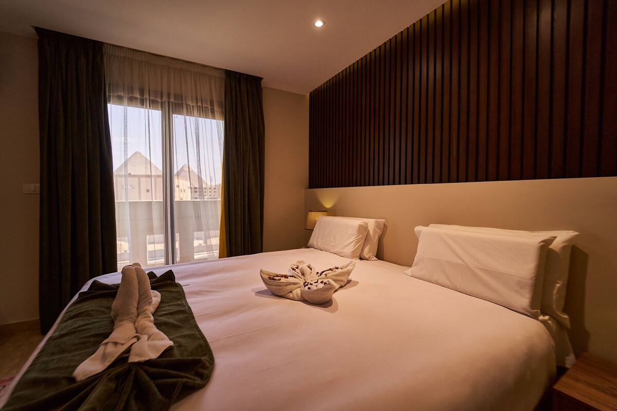 Una habitación de hotel con cama, ventana con cortinas, lámpara de noche y vista a edificios.