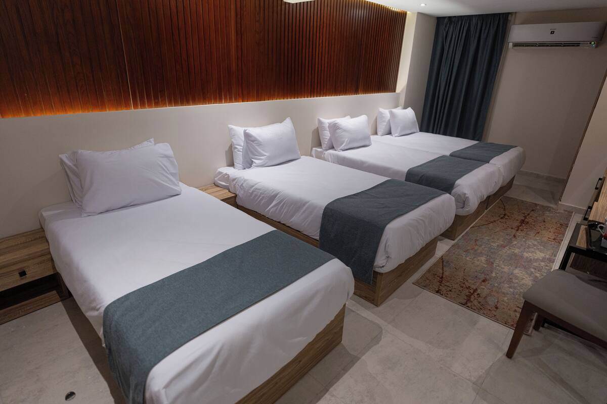 Una habitación de hotel con tres camas, cabecera de madera, un ventanal con cortinas y una pequeña mesa.
