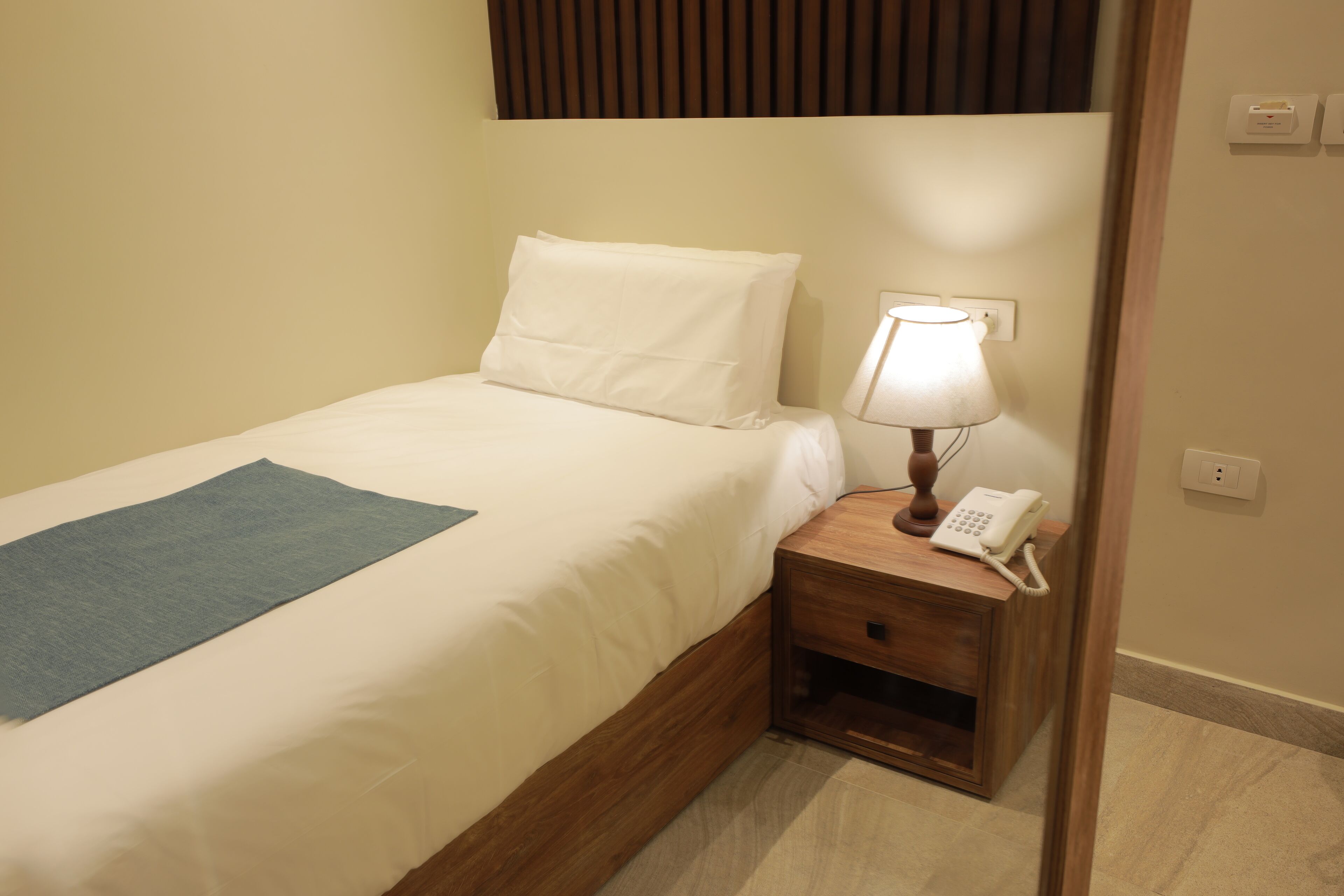 Un lit bien fait, agrémenté d’un couvre-lit bleu, une lampe de chevet et un téléphone posés sur une table de chevet en bois.