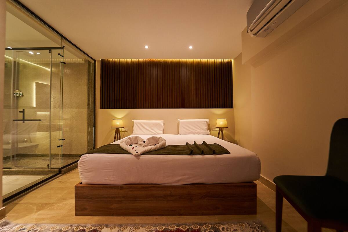 Una habitación de hotel moderna con ducha de vidrio y una cama con plataforma de madera, además de dos lámparas de noche.