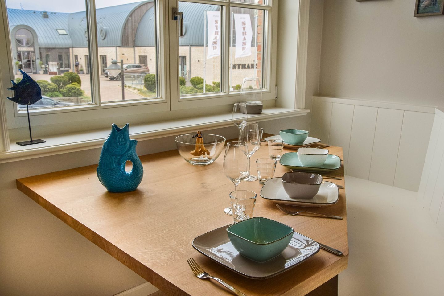 Ein Esstisch mit Geschirr, Gläsern und Schüsseln, eine blaue Vase und ein Blick auf ein Gebäude durch ein Fenster.