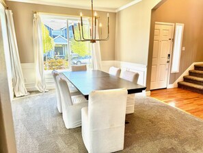 A dining area with a dark wooden table and white upholstered chairs, a chandelier, and a view of a house through a large window with curtains.