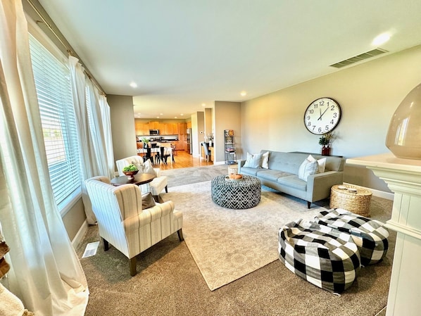 A living room with a sofa, ottoman, and chairs, a kitchen in the background, and a large wall clock.