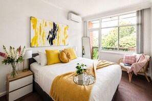 A modern bedroom with a large bed, a bedside table, a window with a view, and a painting on the wall. - The Meadow Gardens Oasis (St Kilda East)