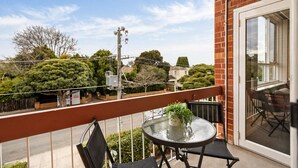 A balcony with a glass table and chairs, a brick wall, and a view of the street and trees. - The Meadow Gardens Oasis (St Kilda East)