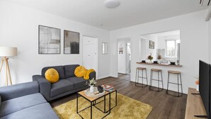 Una sala de estar moderna con un sofá gris, cojines amarillos, una mesa de centro y una zona de cocina con taburetes de bar.