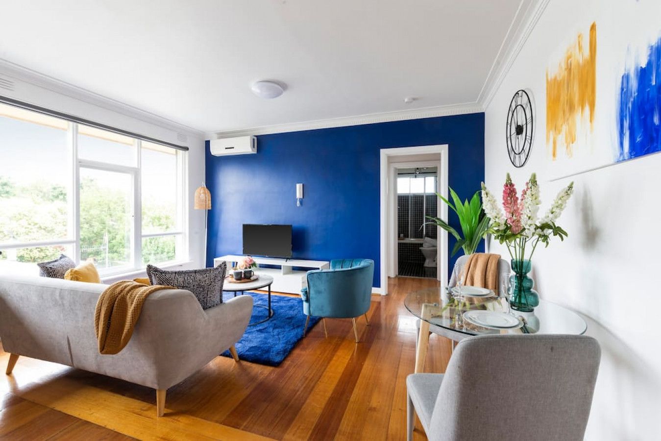 Una sala de estar moderna con una pared azul de acento, un sofá gris, un sillón azul, una mesa de centro de vidrio y un televisor sobre un mueble blanco.