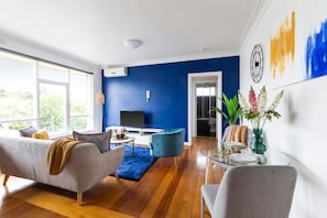 A modern living room with a blue accent wall, a grey sofa, a blue armchair, a glass coffee table, and a TV on a white stand.