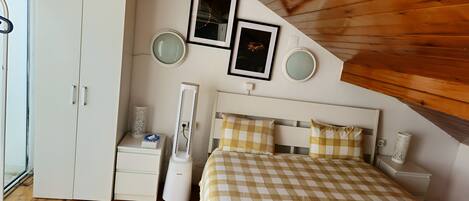 Une chambre avec une couvre-lit à carreaux, une armoire blanche, une table de chevet avec une lampe, et deux tableaux encadrés accrochés au mur.