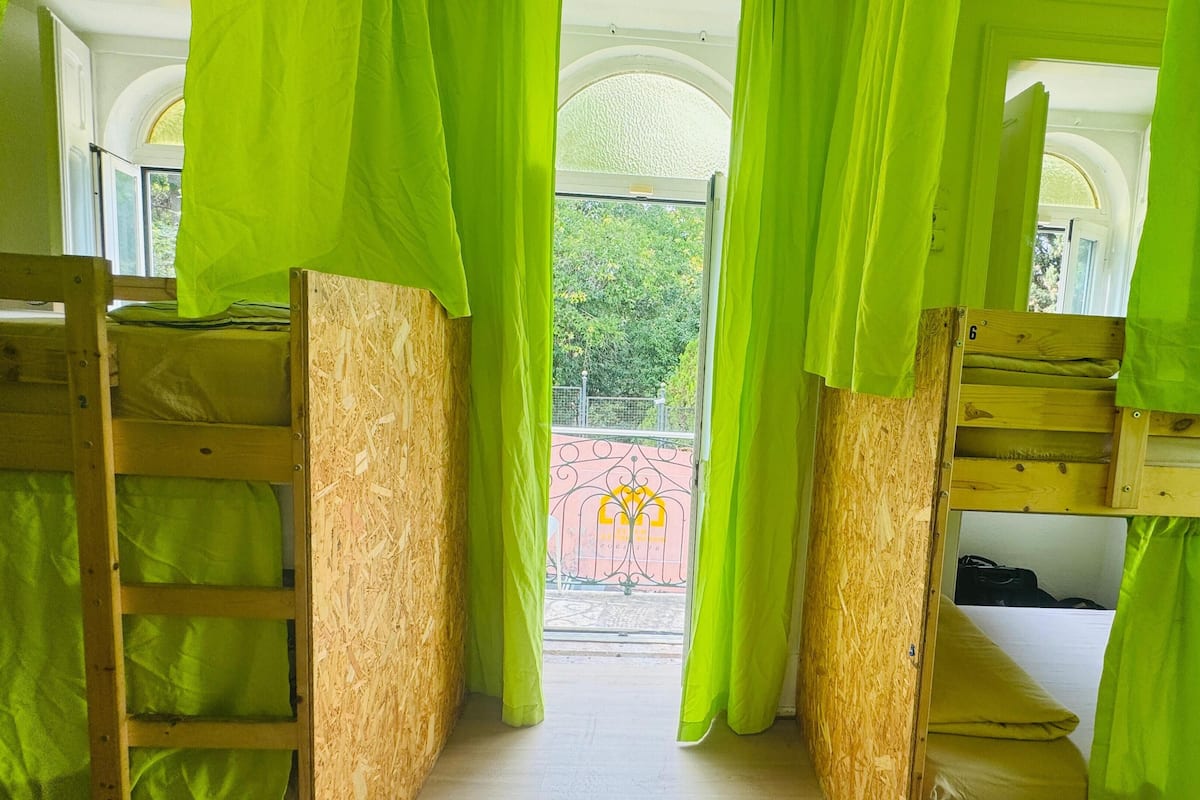 Ein Zimmer mit Etagenbetten, grünen Vorhängen und Blick auf einen Garten durch ein Fenster.