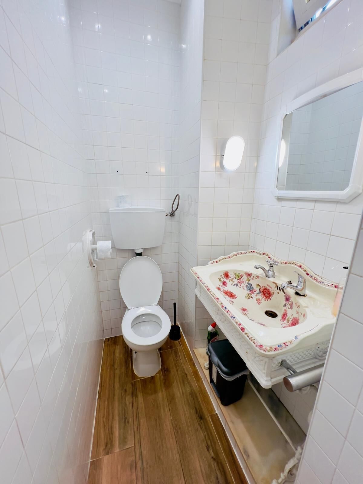 A small, compact bathroom with white tiled walls, a wooden floor, a white toilet, a sink with a floral design, and a mirror above the sink.