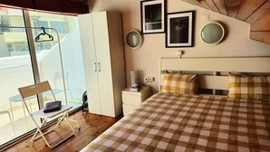 Ein Schlafzimmer mit kariertem Bettbezug, Holzboden, Kleiderschrank, Nachttisch und Stuhl am Fenster.