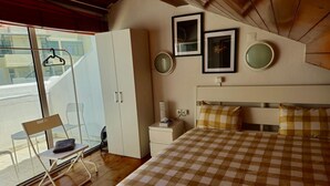 Une chambre avec une couvre-lit à carreaux, une armoire blanche, une table de chevet et une chaise près d’une porte-fenêtre coulissante.