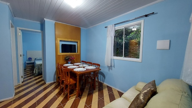 Un comedor con una mesa de madera preparada para seis personas, un televisor montado en la pared y un ventanal con vistas a zonas verdes.