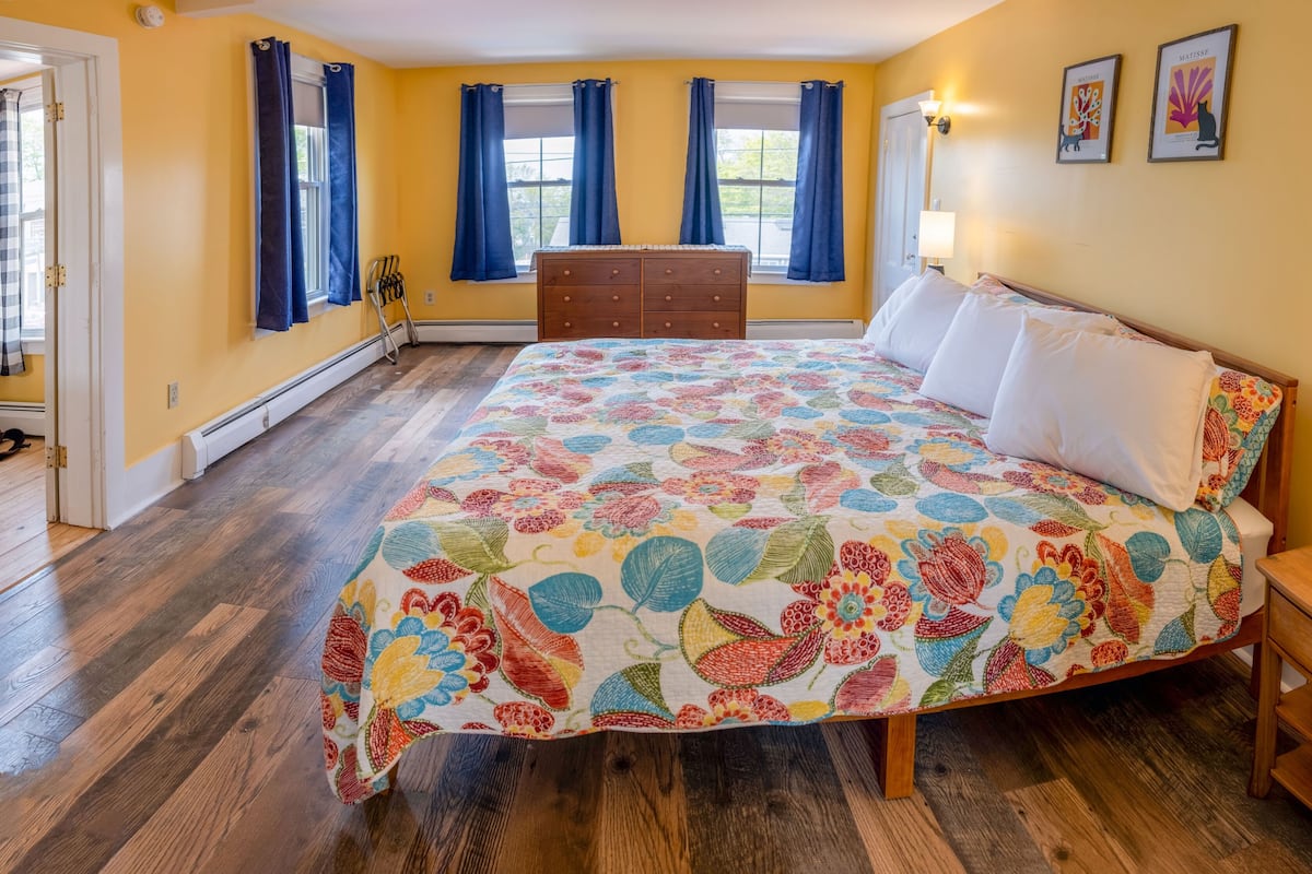 A bedroom with a floral bedspread, wooden dresser, and two windows with blue curtains.