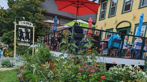 A restaurant with outdoor seating, surrounded by a variety of colorful flowers and a sign advertising breakfast. - Historic 2 Cats Inn (Bar Harbor)