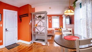 A room with orange walls, a white door, a wooden floor, a dining table with chairs, a hanging lamp, and a white shelving unit with various items. - Historic 2 Cats Inn (Bar Harbor)
