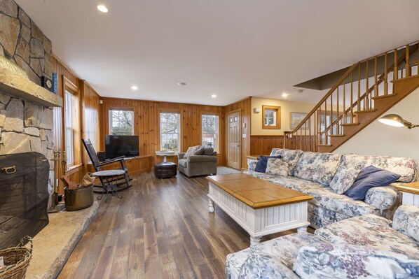 Ein geräumiges Wohnzimmer mit Kamin aus Stein, Holzboden, einem großen Blumensofa, einem Holztisch und einer Treppe.