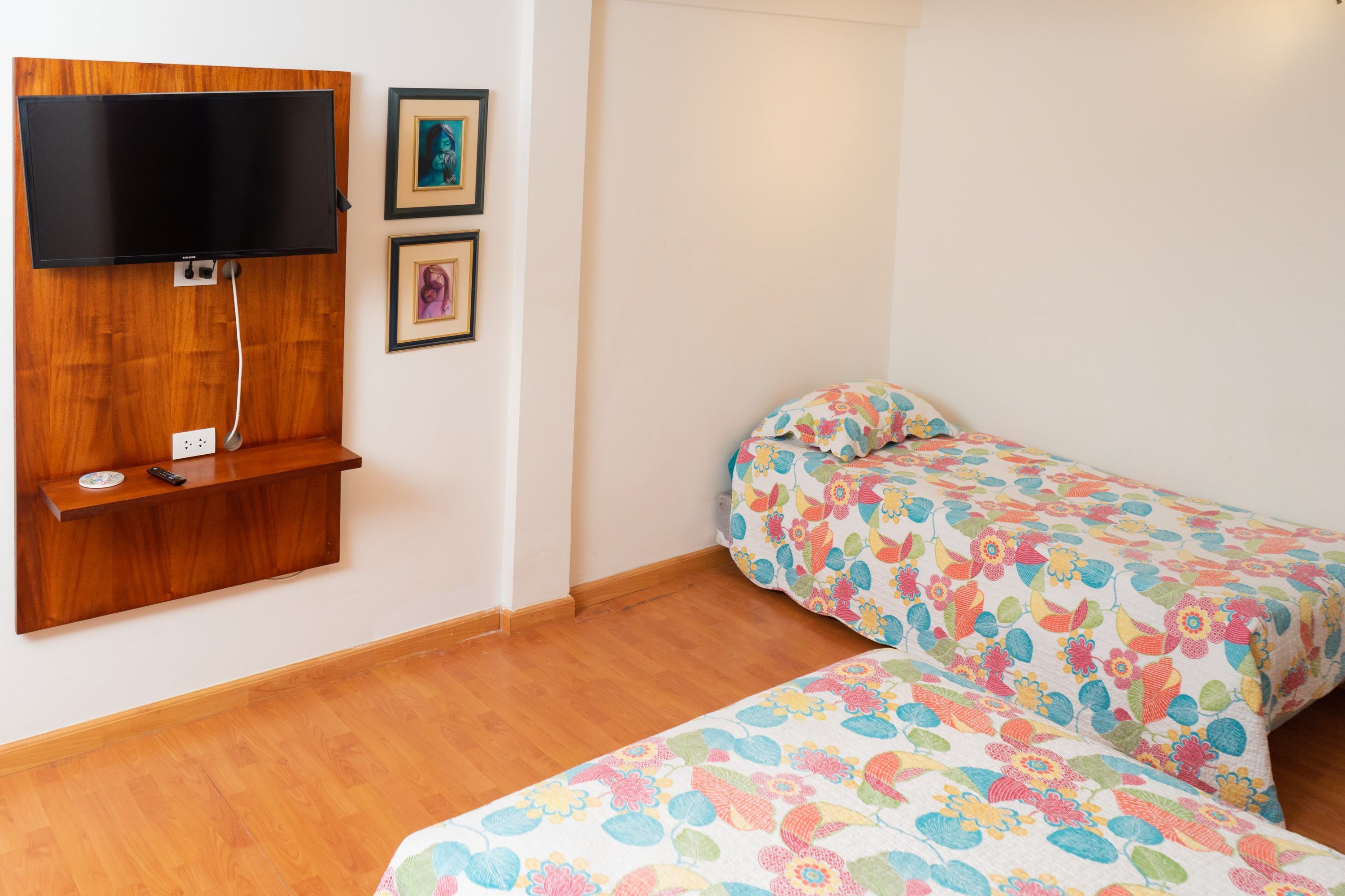 Quarto com duas camas, TV presa em uma prateleira de madeira e parede decorada com dois quadros.