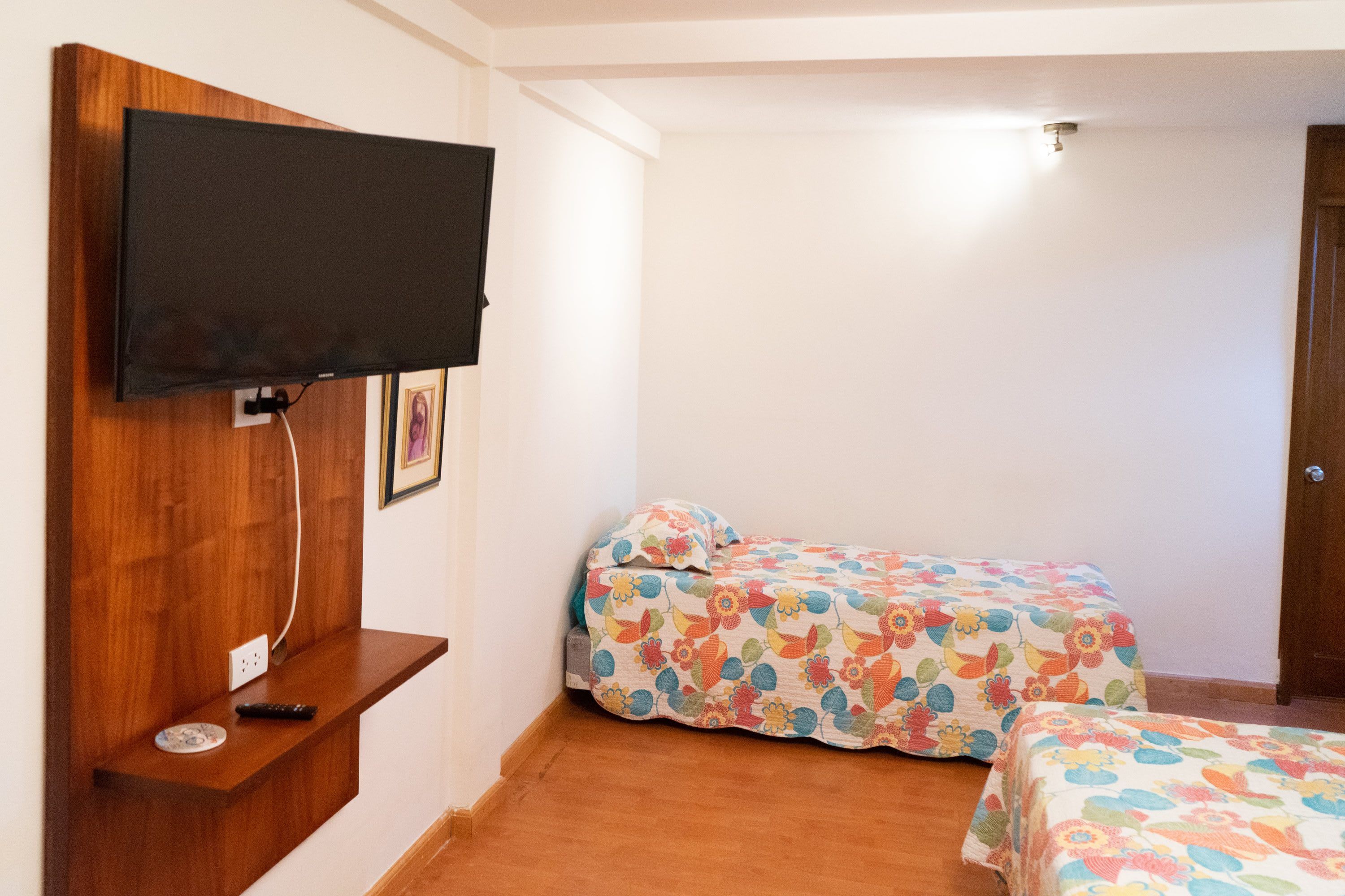 A bedroom with a television mounted on the wall, a bed with floral bedding, and a bedside table with a remote control.