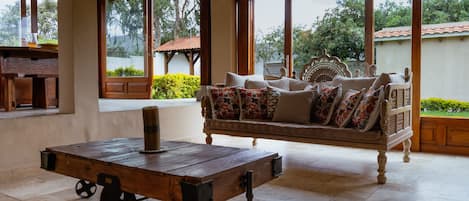 A spacious room with a wooden table, a sofa with patterned cushions, and large windows offering a view of greenery and a building with a red roof.