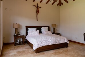 A bedroom with a wooden bed, two bedside tables with lamps, a wall-mounted cross, and a tiled floor. - Guayllabamba Estate - Indoor Pool, Jacuzzi & Gym (Quito)
