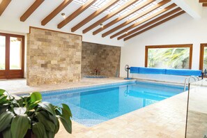Une piscine avec un sol carrelé, des poutres en bois au plafond, un mur en pierre et une fenêtre donnant sur des palmiers.