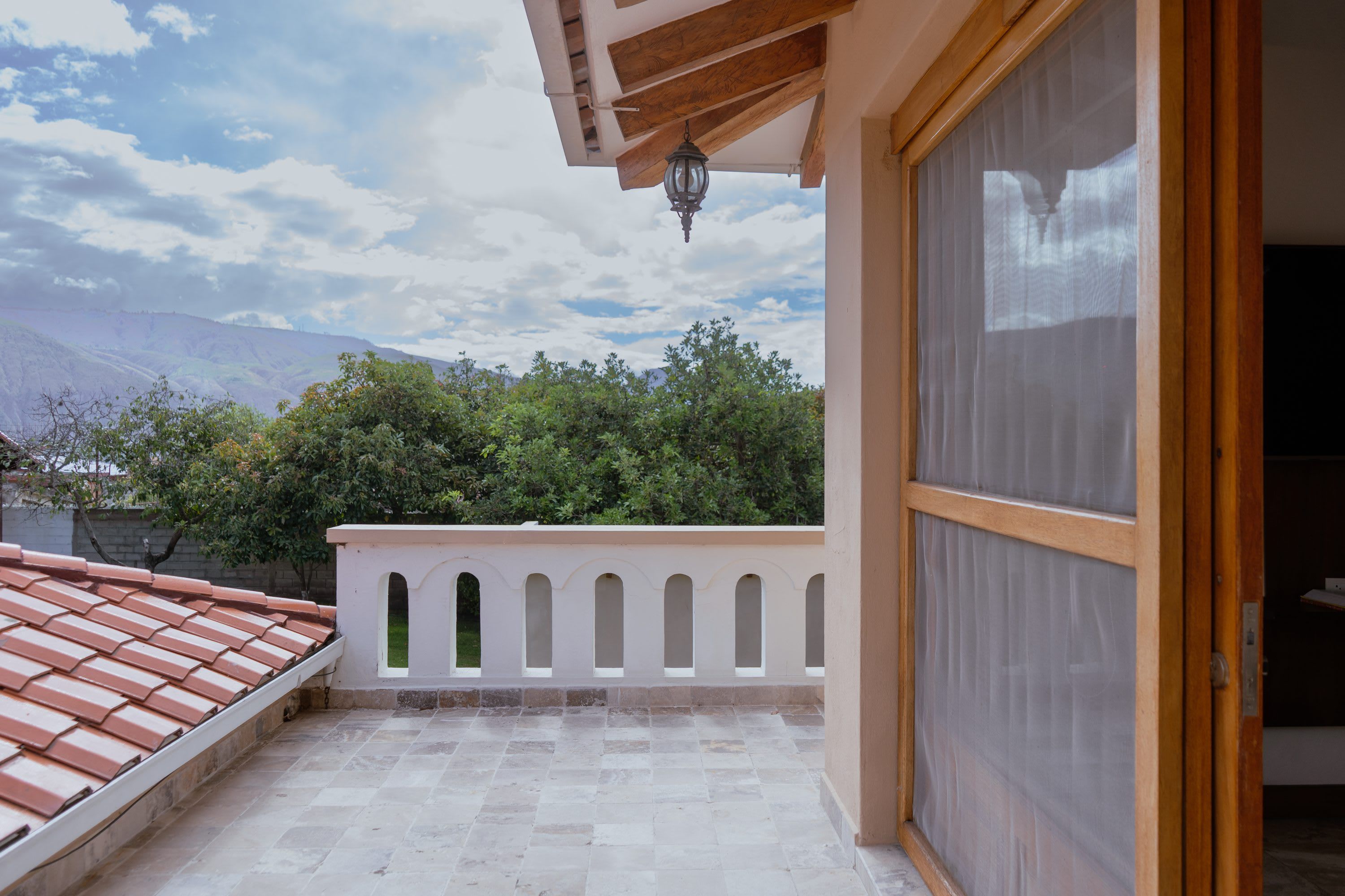 Uma varanda com piso de azulejos, guarda-corpo branco com arcos e vista para árvores e montanhas ao fundo.