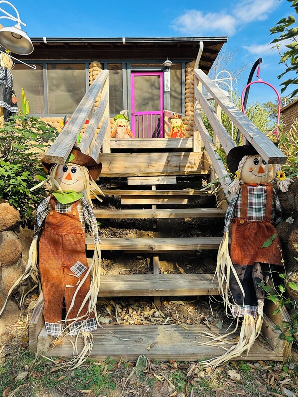 A rustic wooden staircase leading to a house with a pink door, surrounded by greenery and decorative elements.