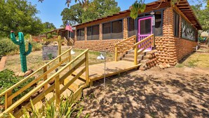 A rustic lodge with a wooden staircase leading to a porch, surrounded by desert vegetation and a cactus.