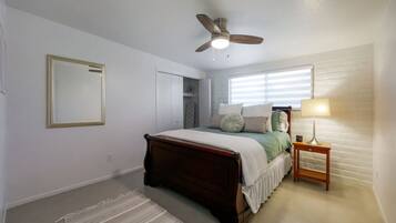 A bedroom with a bed, a ceiling fan, a bedside table with a lamp, a window with blinds, and a door.