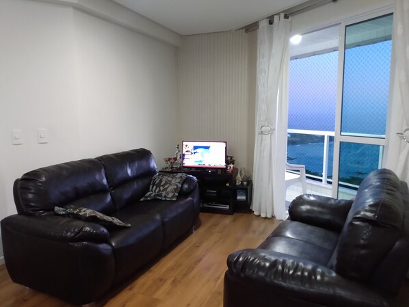 A living room with a black leather sofa, a television, and a view of the sea through large windows.