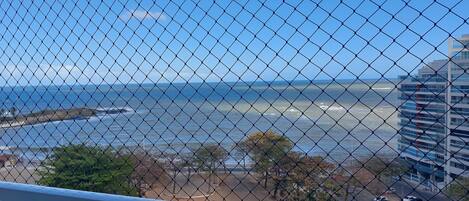 A view through a chain-link fence of a beach with waves, a parking lot, and a building in the background.
