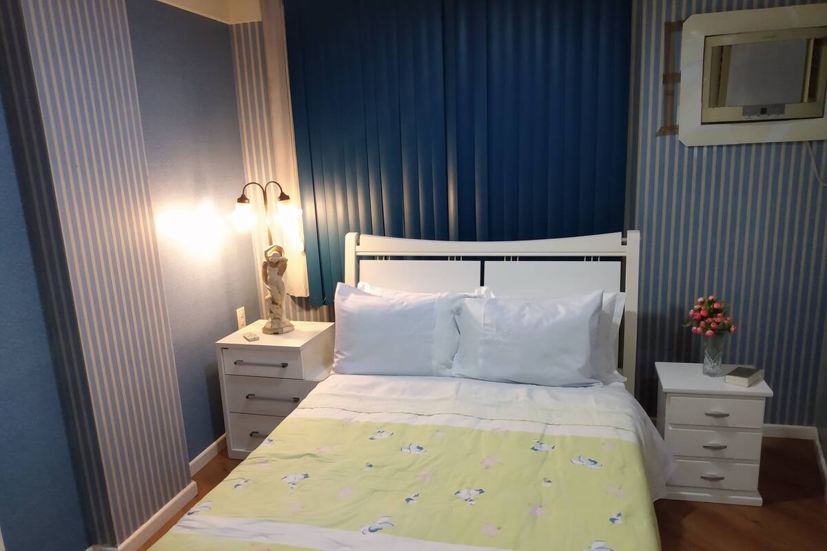 Un lit bien fait, recouvert d’une couvre-lit à motifs floraux, une lampe de chevet et une table de chevet ornée d’un vase de fleurs.