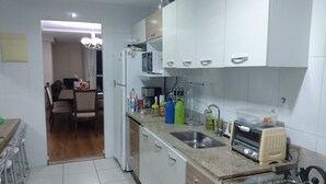 Une cuisine compacte avec des meubles blancs, une cuisinière à gaz, un four à micro-ondes et un évier. On aperçoit un coin repas à l’arrière-plan.