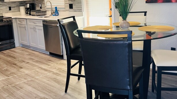A modern kitchen with white cabinets, a built-in oven, and a coffee machine. There is a dining area with a round table and black chairs.