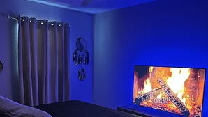 A bedroom with a bed, a ceiling fan, a wall-mounted TV displaying a fireplace, and a dreamcatcher decoration.